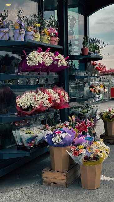 A display of fresh floral arrangements, including colorful bouquets of white, pink, and purple flowers wrapped in vibrant paper and ribbon, arranged on metal shelves inside a flower shop with large gl