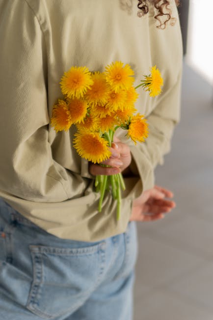 A person wearing a beige shirt and light blue jeans holding a bouquet of bright yellow carnations with lush, ruffled petals and green stems. The bouquet is gathered in the person's left hand, with the