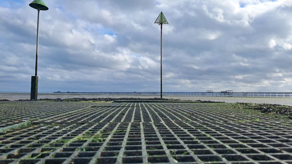 A wide-angle view of a seaside promenade featuring two tall, thin street lamps with minimalist green metal tops. The foreground shows a textured, grid-like surface made of stone tiles with small patch